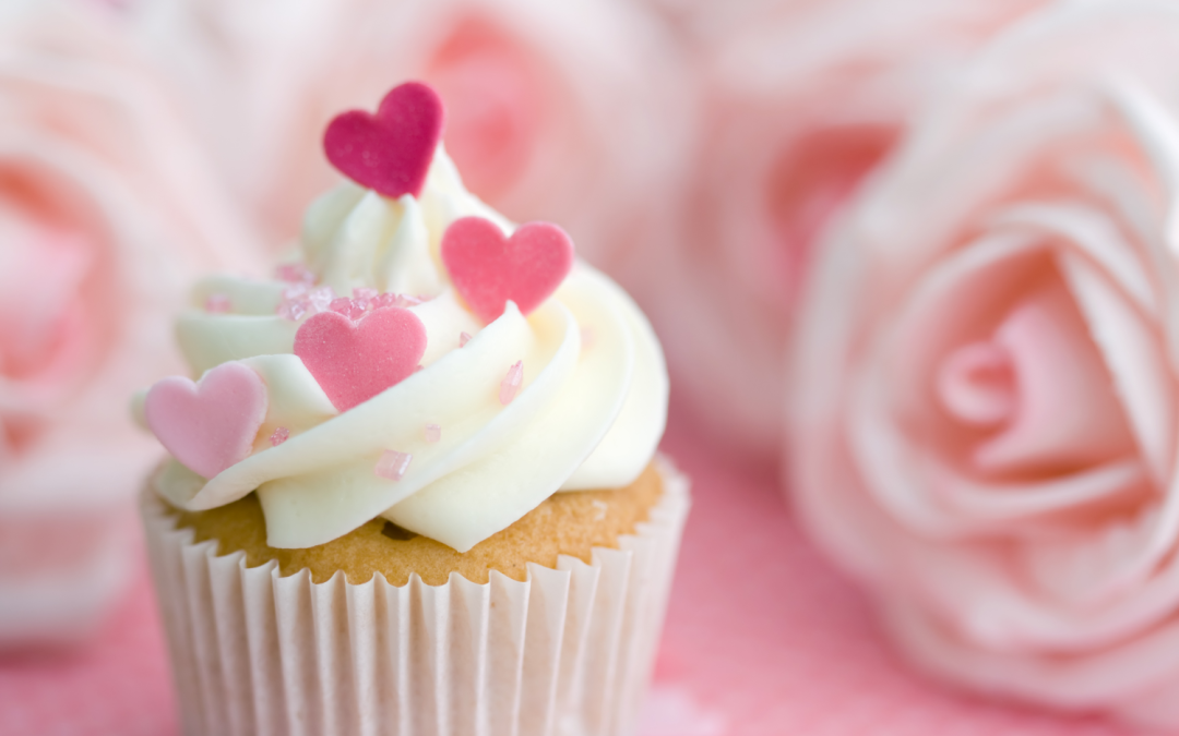 cupcake décoré de crème onctueuse et de petits cœurs roses et rouges, accompagné de sucre scintillant, posé sur une surface rose avec des roses en arrière-plan. Une ambiance douce et romantique, parfaite pour une célébration de Galentine’s Day, entre amies, avec des douceurs et une décoration élégante.